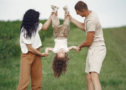 A joyful family enjoys outdoor play, lifting their child upside down in a green field.